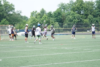 A group of athletes playing lacrosse on a field. They are wearing helmets and various sports gear. One player is jumping in the air, holding a lacrosse stick, while others are positioned across the field, some wearing numbered jerseys. Trees and a netted goal are visible in the background, providing a natural backdrop.