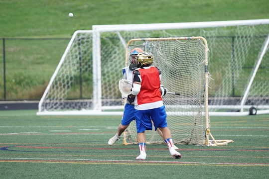 Two lacrosse players wearing protective gear are actively engaged in the game on a field. One player, in a red vest and helmet, is facing the goal and appears to be attempting a shot, while the other player, wearing blue shorts and protective equipment, is positioned as the goalkeeper. The outdoor sports field is marked with lines and surrounded by green grass and a fence.