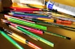 A variety of art supplies are scattered across a yellow surface, including paintbrushes, pencils, and paint tubes. The brushes and pencils are in vibrant colors such as red, green, and purple, while the paint tubes are predominantly white and blue.