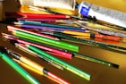 A variety of art supplies are scattered across a yellow surface, including paintbrushes, pencils, and paint tubes. The brushes and pencils are in vibrant colors such as red, green, and purple, while the paint tubes are predominantly white and blue.