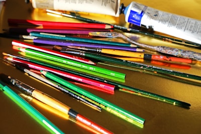 A variety of art supplies are scattered across a yellow surface, including paintbrushes, pencils, and paint tubes. The brushes and pencils are in vibrant colors such as red, green, and purple, while the paint tubes are predominantly white and blue.