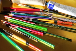 A variety of art supplies are scattered across a yellow surface, including paintbrushes, pencils, and paint tubes. The brushes and pencils are in vibrant colors such as red, green, and purple, while the paint tubes are predominantly white and blue.