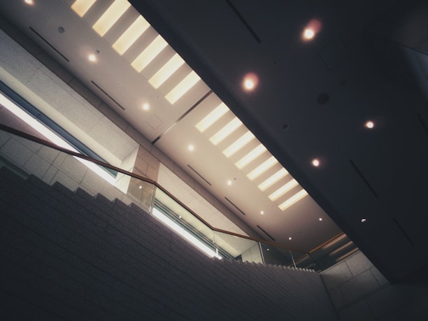 An interior space with a modern staircase leading upwards, featuring sleek metal and glass railings. The ceiling displays a series of evenly spaced, rectangular light fixtures that provide a soft, warm glow. The architecture emphasizes clean lines and minimalist design, with concrete or stone walls adding to the contemporary aesthetic.