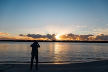 A person stands on a beach capturing a photograph of a vibrant sunset. The sun is setting behind a line of dark, silhouetted clouds and trees on the horizon. The sky is painted with shades of orange, yellow, and blue, reflected on the calm water surface.