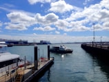 A marina with several boats is docked alongside the pier. A large cargo ship bearing the logo 'NYK Line' is anchored in the distance. A smaller vessel appears to be navigating the waterway toward the port. The sky is mostly clear, with scattered white clouds and ample sunlight reflecting off the water.