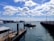 A marina with several boats is docked alongside the pier. A large cargo ship bearing the logo 'NYK Line' is anchored in the distance. A smaller vessel appears to be navigating the waterway toward the port. The sky is mostly clear, with scattered white clouds and ample sunlight reflecting off the water.