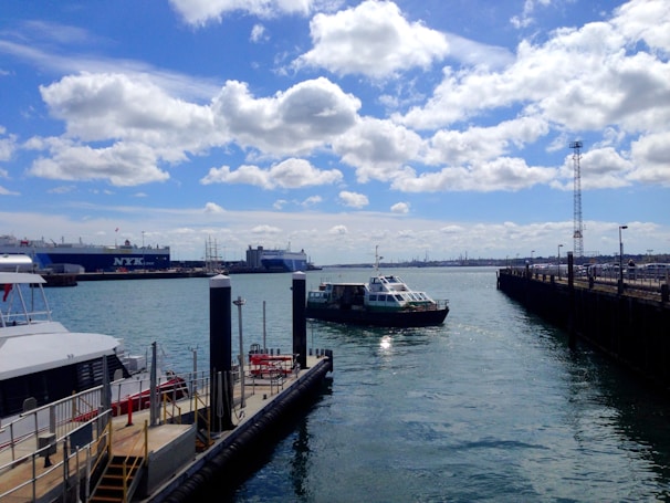 A marina with several boats is docked alongside the pier. A large cargo ship bearing the logo 'NYK Line' is anchored in the distance. A smaller vessel appears to be navigating the waterway toward the port. The sky is mostly clear, with scattered white clouds and ample sunlight reflecting off the water.