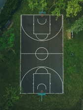 An aerial view of a basketball court surrounded by lush green grass. The court features standard markings with two basketball hoops, and there is a bench nearby. Trees partially overhang the court, creating a serene outdoor environment.