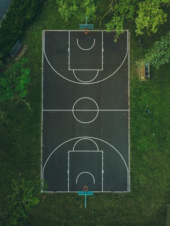 An aerial view of a basketball court surrounded by lush green grass. The court features standard markings with two basketball hoops, and there is a bench nearby. Trees partially overhang the court, creating a serene outdoor environment.