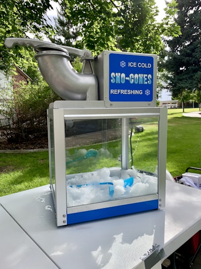 A sno-cone machine is situated on a white table outdoors. The machine has a metallic top with a large spout and a clear glass compartment filled with ice. A blue sign on the top reads 'ICE COLD SNO-CONES REFRESHING' with snowflake symbols. The background has lush green trees and a well-maintained lawn.