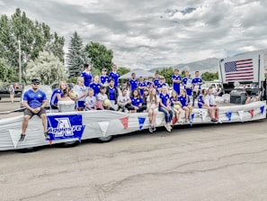 A group of young people and adults, dressed in matching blue sports jerseys, are sitting and standing on a decorated parade float. Some are holding soccer balls and an American flag is prominently displayed. The setting appears to be a parade with trees and cars in the background.