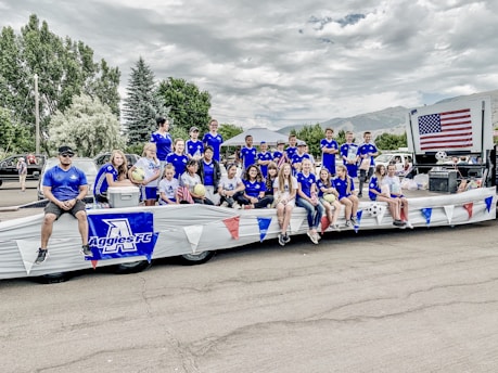 A group of young people and adults, dressed in matching blue sports jerseys, are sitting and standing on a decorated parade float. Some are holding soccer balls and an American flag is prominently displayed. The setting appears to be a parade with trees and cars in the background.