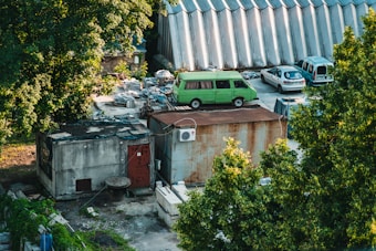 The image includes an industrial area with a rusted metal structure and a green van parked nearby. The setting is surrounded by lush green trees, adding contrast to the industrial feel. Several other vehicles are parked near stacks of construction materials, and a corrugated metal building is visible in the background.