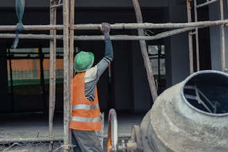 A construction worker wearing a green helmet and an orange safety vest is adjusting metal scaffolding. The environment suggests an active construction site with unfinished structures. A concrete mixer is visible in the foreground on the right side.