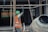 A construction worker wearing a green helmet and an orange safety vest is adjusting metal scaffolding. The environment suggests an active construction site with unfinished structures. A concrete mixer is visible in the foreground on the right side.