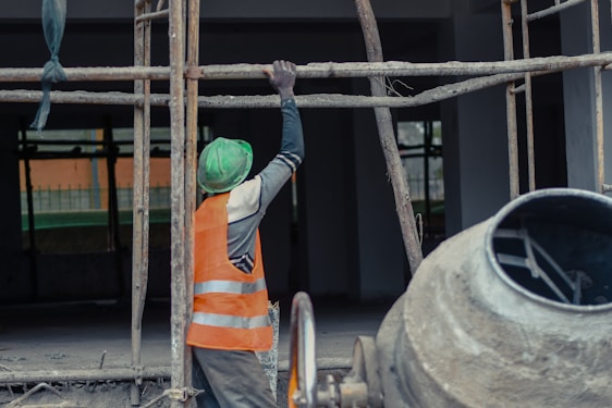 A construction worker wearing a green helmet and an orange safety vest is adjusting metal scaffolding. The environment suggests an active construction site with unfinished structures. A concrete mixer is visible in the foreground on the right side.