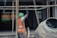 A construction worker wearing a green helmet and an orange safety vest is adjusting metal scaffolding. The environment suggests an active construction site with unfinished structures. A concrete mixer is visible in the foreground on the right side.