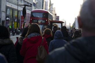 A bustling city street with people walking in various directions. A red double-decker bus, typical of London, is prominently visible. The surrounding buildings are urban and commercial, with various shop signs and advertisements. The lighting suggests early evening, casting long shadows.