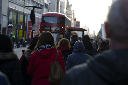 A bustling city street with people walking in various directions. A red double-decker bus, typical of London, is prominently visible. The surrounding buildings are urban and commercial, with various shop signs and advertisements. The lighting suggests early evening, casting long shadows.