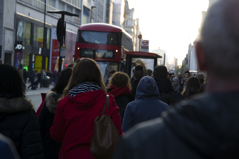 A bustling city street with people walking in various directions. A red double-decker bus, typical of London, is prominently visible. The surrounding buildings are urban and commercial, with various shop signs and advertisements. The lighting suggests early evening, casting long shadows.