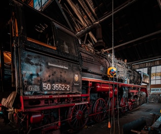 Inside an old rail workshop, a vintage steam locomotive is prominently displayed. The locomotive has a dark metal body with red accents, featuring large wheels and intricate mechanical details. The environment suggests an industrial setting with metal beams and glass windows allowing natural light to enter.