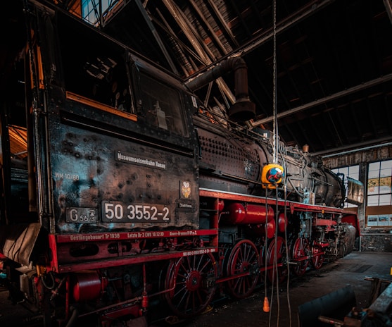 Inside an old rail workshop, a vintage steam locomotive is prominently displayed. The locomotive has a dark metal body with red accents, featuring large wheels and intricate mechanical details. The environment suggests an industrial setting with metal beams and glass windows allowing natural light to enter.