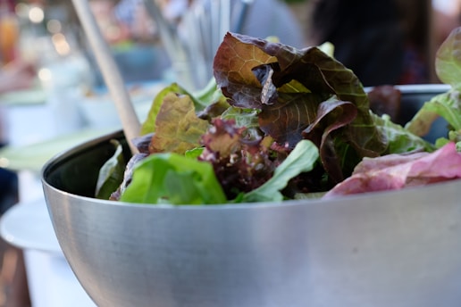A close-up view of a fresh salad with a mix of green and red leafy lettuce in a metallic bowl. Wooden salad tongs and a blurred background suggest a casual outdoor dining setting.