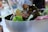 A close-up view of a fresh salad with a mix of green and red leafy lettuce in a metallic bowl. Wooden salad tongs and a blurred background suggest a casual outdoor dining setting.