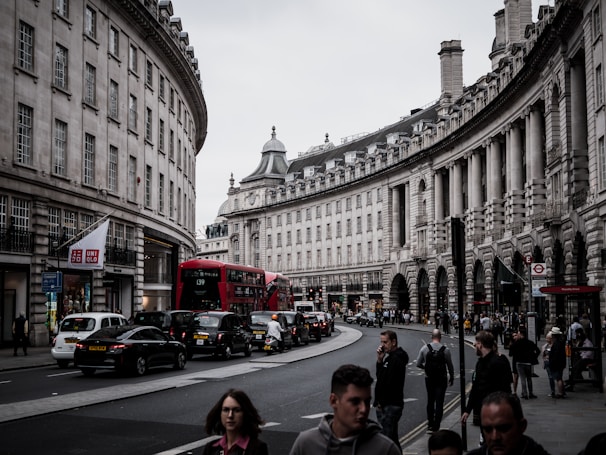 A busy urban street scene featuring a double-decker red bus moving alongside a row of black cabs and other vehicles. The architecture includes classic, curved stone buildings with many windows and ornate detailing. Pedestrians are walking along the sidewalks, and there's a store with a visible signage for Uniqlo.