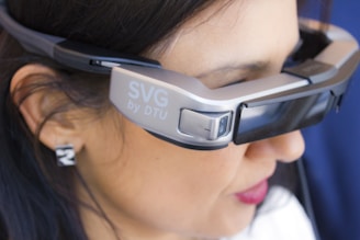 A person is wearing a pair of advanced augmented reality glasses with the label 'SVG by DTU' on the side, indicating a technological product. The glasses have a sleek, modern design and cover the eyes of the individual. The person has dark hair and is seen in a close-up view, focusing primarily on the device.