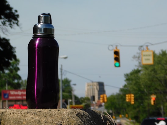 A dark purple metallic water bottle with a silver cap sits on a concrete surface. In the background, a traffic light is visible showing green, and trees line the sides of a city street. Buildings can be seen in the distance under a clear sky.