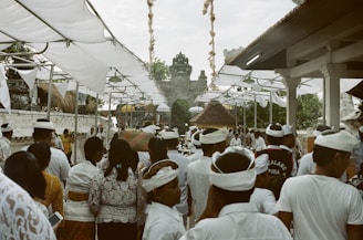 A large group of people dressed in traditional white attire gather under a canopy, which is decorated with hanging fabric. The setting appears to be a cultural or religious event with a stone temple gate visible in the background.