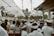 A large group of people dressed in traditional white attire gather under a canopy, which is decorated with hanging fabric. The setting appears to be a cultural or religious event with a stone temple gate visible in the background.