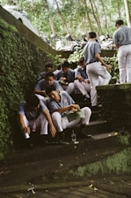 A group of men in matching uniforms sit on stone steps surrounded by lush greenery. Some men are eating from green leaves while others are engaged in conversation. Motorbikes and helmets are parked above the steps. The environment is outdoors, possibly in a shaded area under trees.