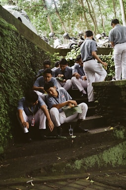 A group of men in matching uniforms sit on stone steps surrounded by lush greenery. Some men are eating from green leaves while others are engaged in conversation. Motorbikes and helmets are parked above the steps. The environment is outdoors, possibly in a shaded area under trees.