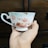 A delicate teacup with floral patterns in shades of pink and red is held by a hand. The background features a dark wooden surface, possibly furniture.