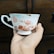 A delicate teacup with floral patterns in shades of pink and red is held by a hand. The background features a dark wooden surface, possibly furniture.