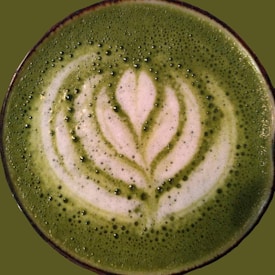 A cup filled with matcha latte, decorated with intricate latte art that resembles a leaf. The surface is smooth with tiny bubbles, and the art is prominently visible in white against the deep green background.
