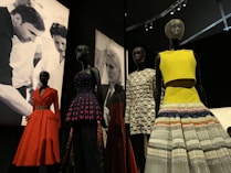 Several mannequins display a variety of colorful and elegant dresses, including a red coat, a dress with a purple and black pattern, and a yellow top with a multicolored skirt. The backdrop features black-and-white images of people engaged in design or fashion activities.