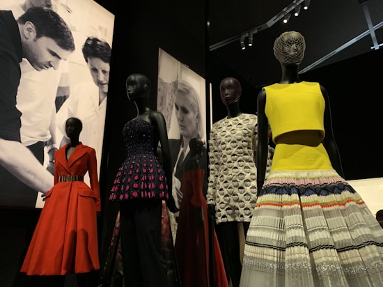 Several mannequins display a variety of colorful and elegant dresses, including a red coat, a dress with a purple and black pattern, and a yellow top with a multicolored skirt. The backdrop features black-and-white images of people engaged in design or fashion activities.