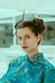 A person is wearing a traditional blue garment with intricate floral and dragon patterns. The hair is styled in two buns with chopsticks inserted for decoration. The background is softly blurred, emphasizing the person.