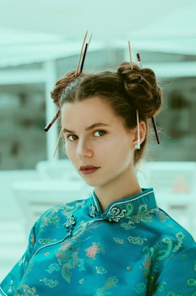A person is wearing a traditional blue garment with intricate floral and dragon patterns. The hair is styled in two buns with chopsticks inserted for decoration. The background is softly blurred, emphasizing the person.