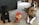 Two dogs sit on a carpeted floor next to a large container of dog treats. One dog is small and dark-colored, while the other is larger with a brindle coat. The container is clear with a red lid, filled with treats. The room contains various objects, including a dog toy and furniture.