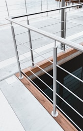 A modern railing with a sleek, metallic design overlooks a body of water. The structure is composed of evenly spaced metal bars and wooden accents, providing a contemporary aesthetic to the environment. The space around the railing features a concrete platform and hints at a possible dock or waterfront setting.