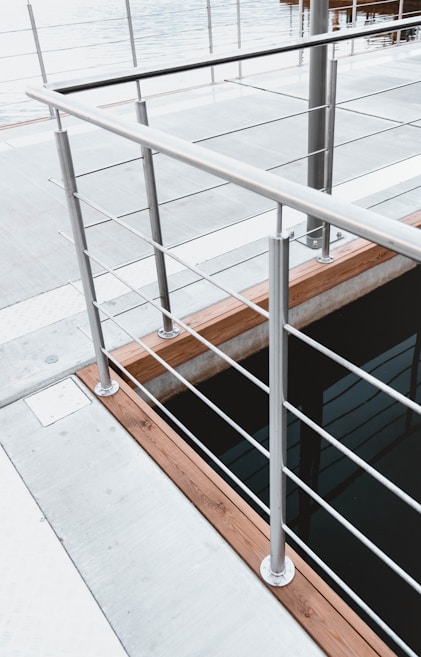A modern railing with a sleek, metallic design overlooks a body of water. The structure is composed of evenly spaced metal bars and wooden accents, providing a contemporary aesthetic to the environment. The space around the railing features a concrete platform and hints at a possible dock or waterfront setting.