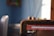 A close-up of a digital device screen showing the Spotify logo and part of a web browser tab with a music playlist. The screen has a noticeable crack on the surface. The background includes a blurred view of a room with a blue wall and possibly a chair or other furniture.