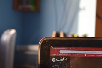A close-up of a digital device screen showing the Spotify logo and part of a web browser tab with a music playlist. The screen has a noticeable crack on the surface. The background includes a blurred view of a room with a blue wall and possibly a chair or other furniture.