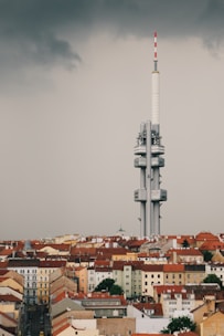 A large, modern telecommunications tower rises above a cityscape filled with older, red-roofed buildings. The sky is overcast, with dark, moody clouds adding a dramatic contrast to the scene.