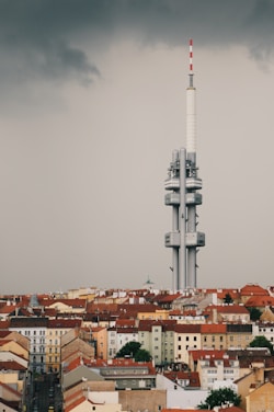 A large, modern telecommunications tower rises above a cityscape filled with older, red-roofed buildings. The sky is overcast, with dark, moody clouds adding a dramatic contrast to the scene.