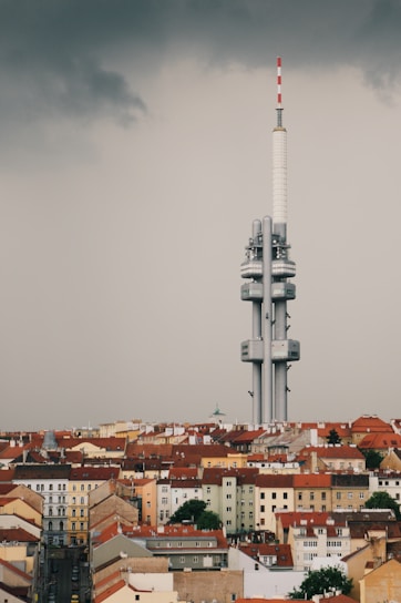 A large, modern telecommunications tower rises above a cityscape filled with older, red-roofed buildings. The sky is overcast, with dark, moody clouds adding a dramatic contrast to the scene.